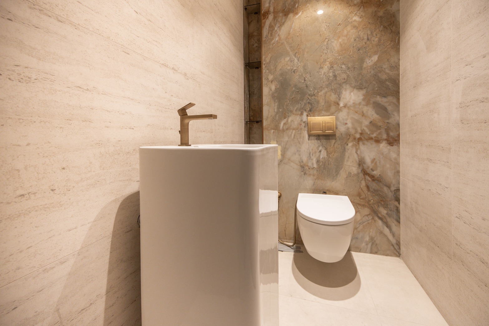 Pearl House By Imtiaz A modern bathroom featuring a white pedestal sink with a gold faucet and a wall-mounted toilet against stone walls.